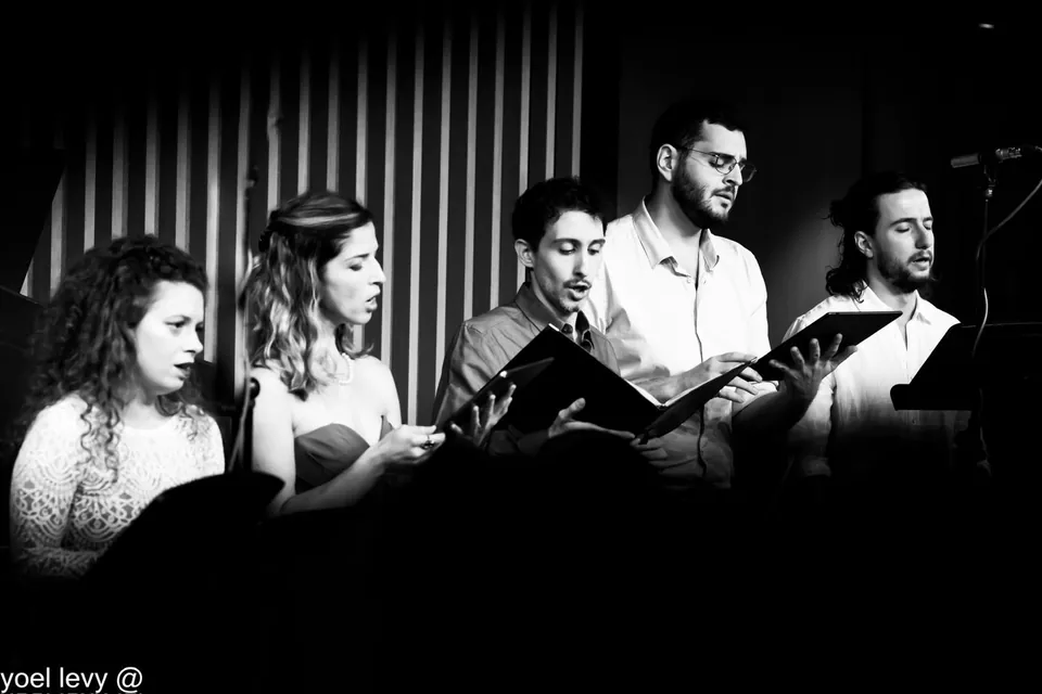 Black-and-white photograph of five singers reading from music folders; a photographer’s watermark is kept at the lower left.