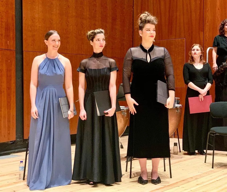Concert-stage photograph of performers in blue and black dresses holding music folders, in front of wood panelling with chairs and percussion behind.